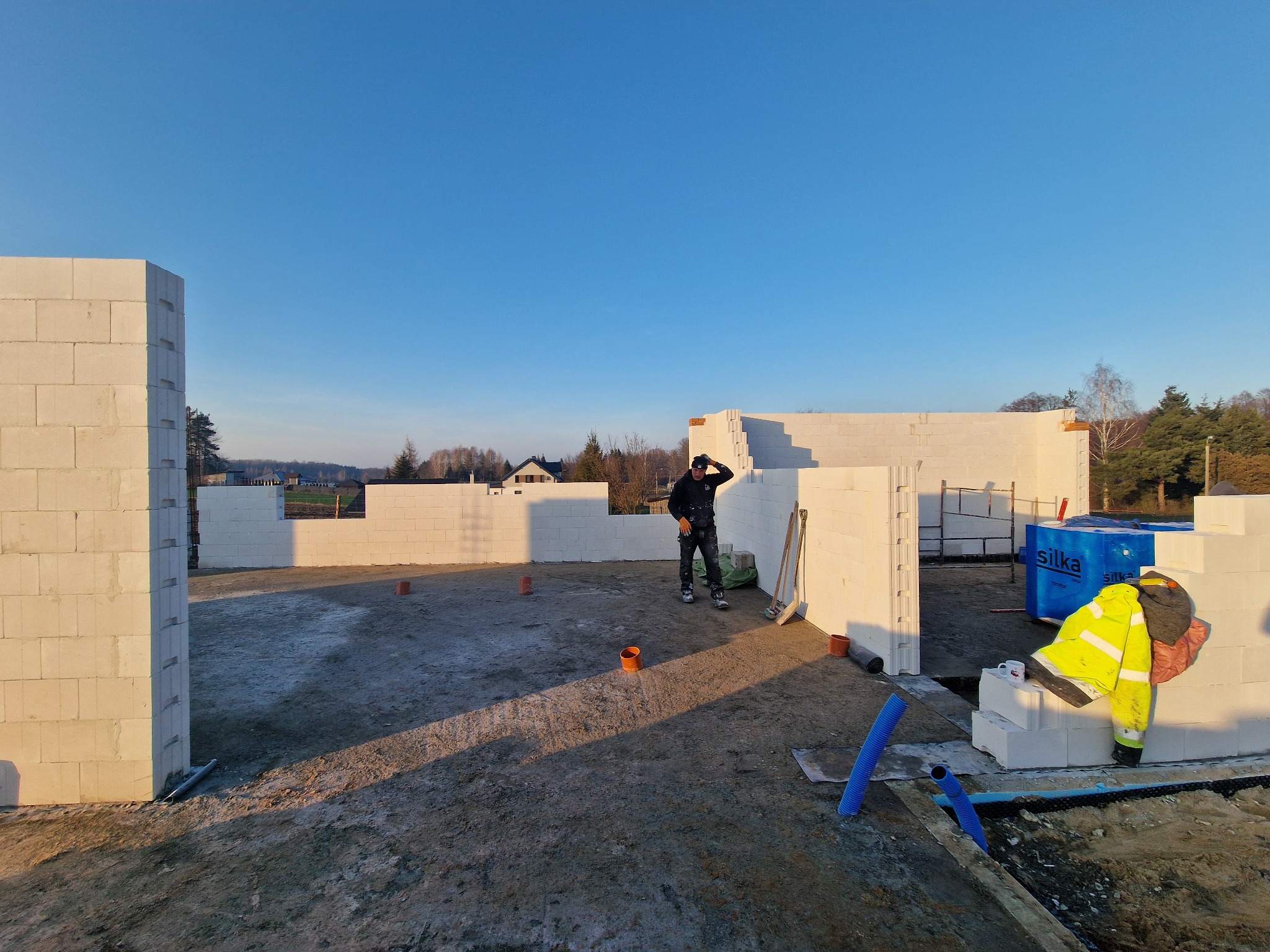 Wznoszenie ścian domu z bloczków silikatowych, widoczny murarz w czarnym ubraniu, narzędzia budowlane, bloczki Silka oraz częściowo ukończona konstrukcja w promieniach zachodzącego słońca.