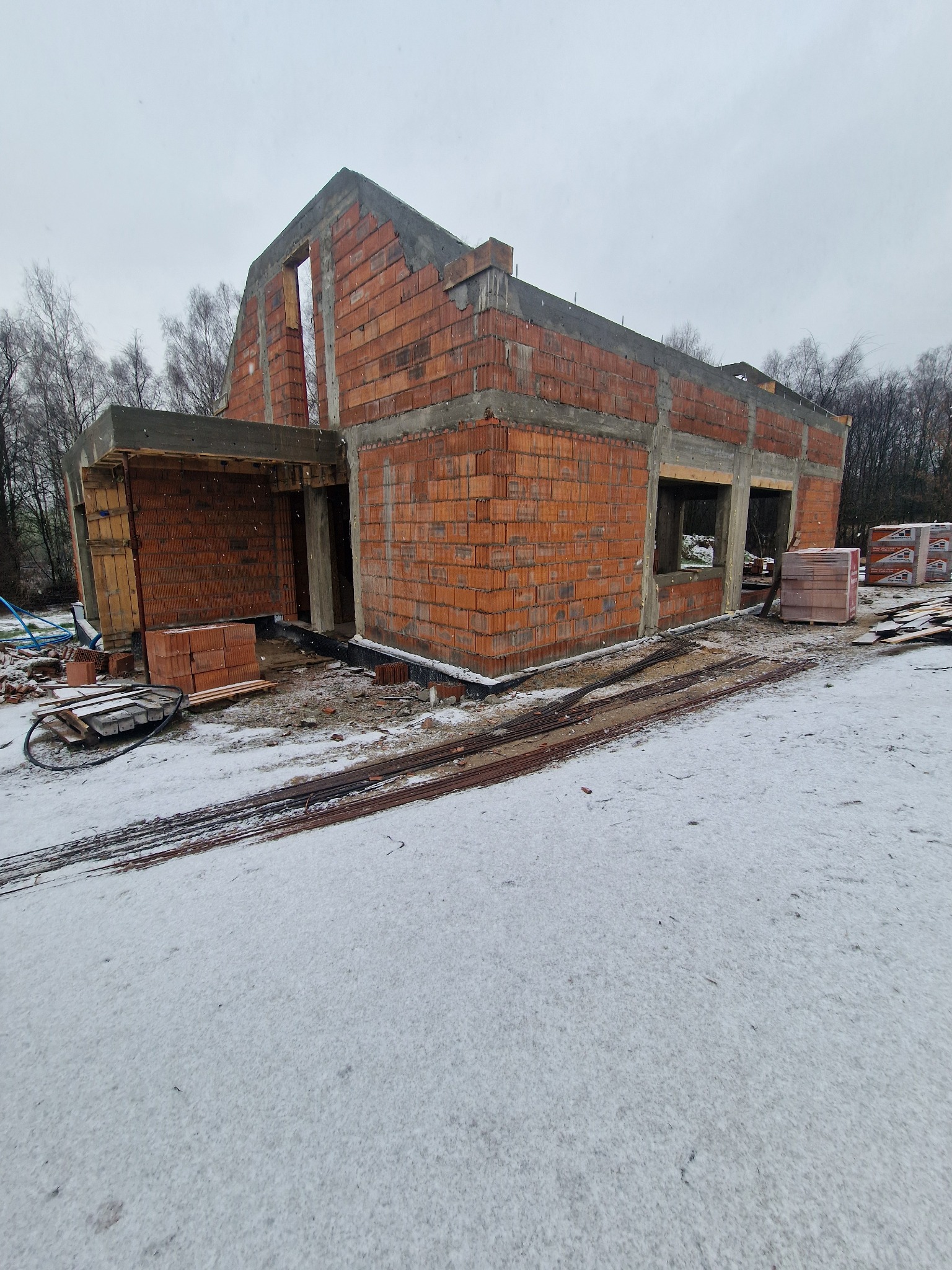Surowy stan murowanego domu z czerwonej cegły w zimowej scenerii, widoczne zbrojenia i palety cegieł, etap budowy.