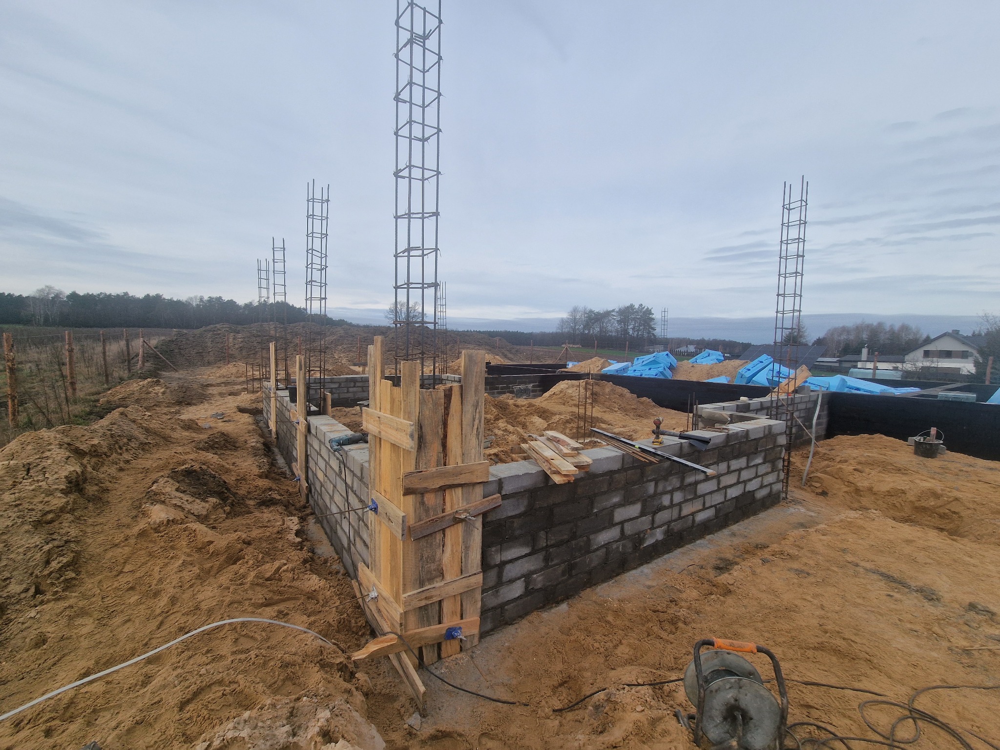 Budowa fundamentów domu z bloczków betonowych, widoczne zbrojenia kolumn, drewniane szalunki narożne, teren budowy zasypany piaskiem, w tle drzewa i budynki.