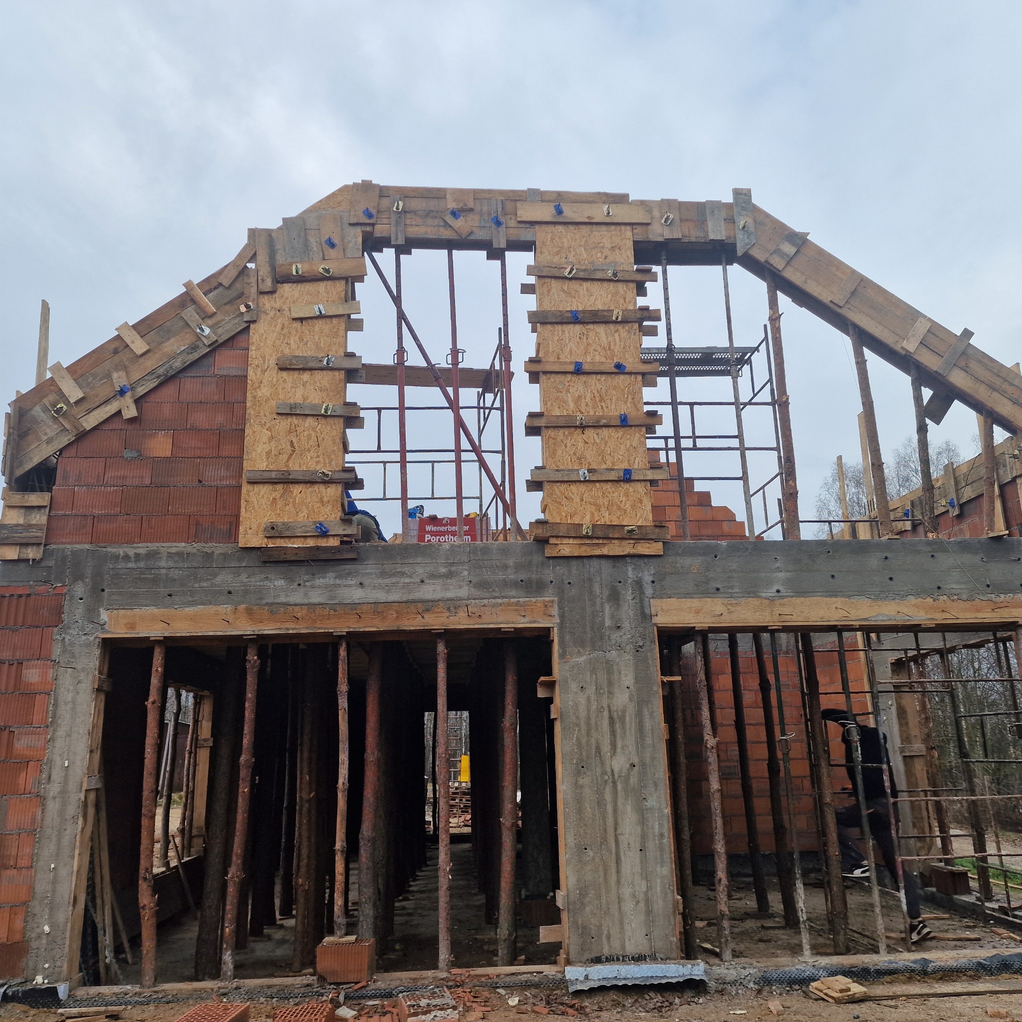 Budowa domu w Łodzi w trakcie wznoszenia ścian z czerwonej cegły Porotherm, widoczne szalunki drewniane pod wylewkę stropu i konstrukcję dachu.