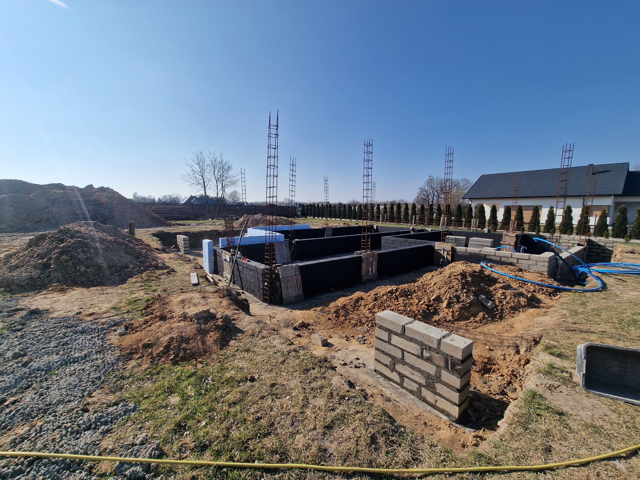 Fundamenty domu w trakcie budowy, widoczne zbrojenia, bloczki betonowe, izolacja termiczna i wykopana ziemia na tle błękitnego nieba.