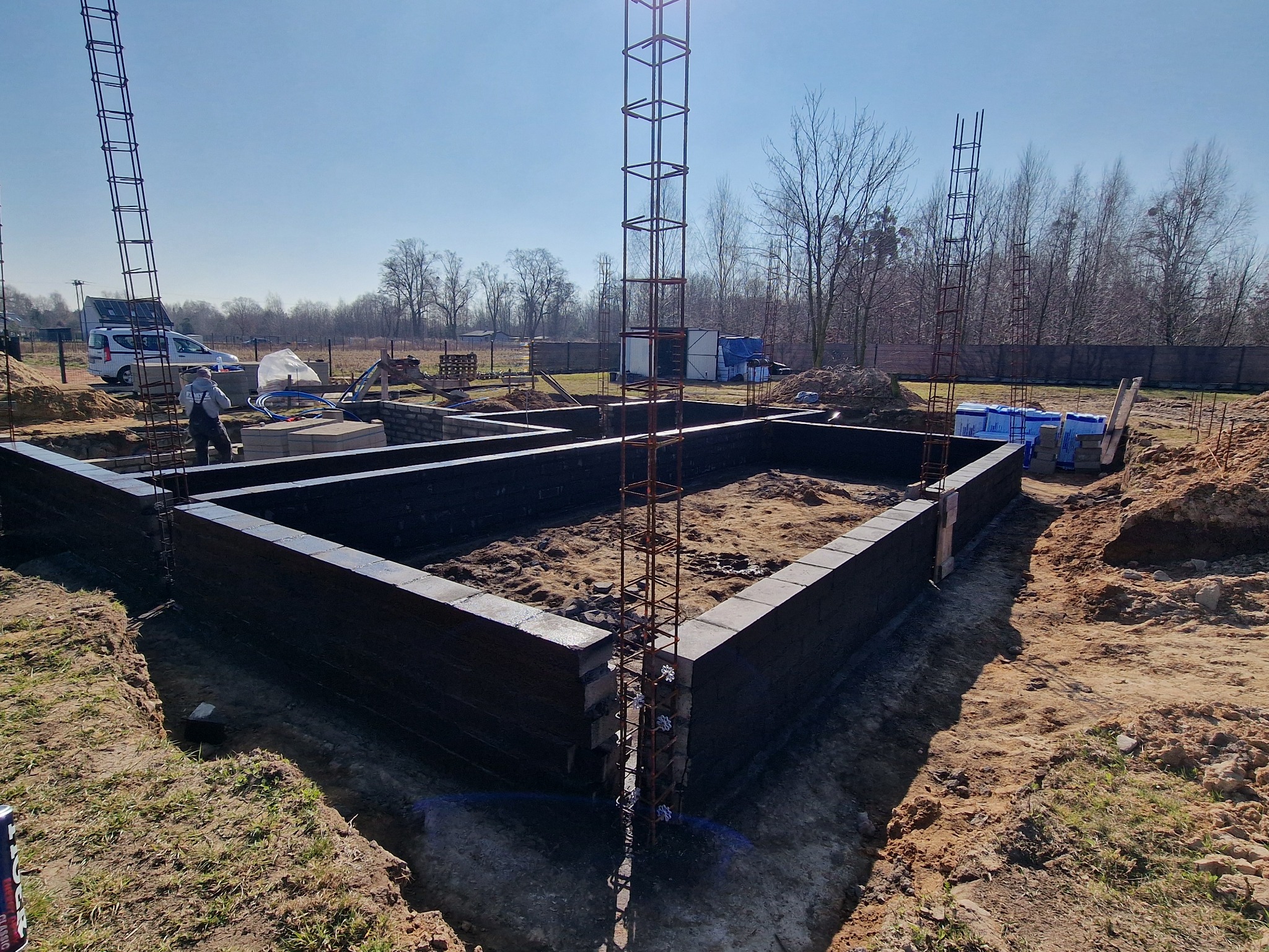 Wykop pod fundamenty z ułożonymi bloczkami betonowymi i zbrojeniem pionowym, widoczny pracownik w tle.