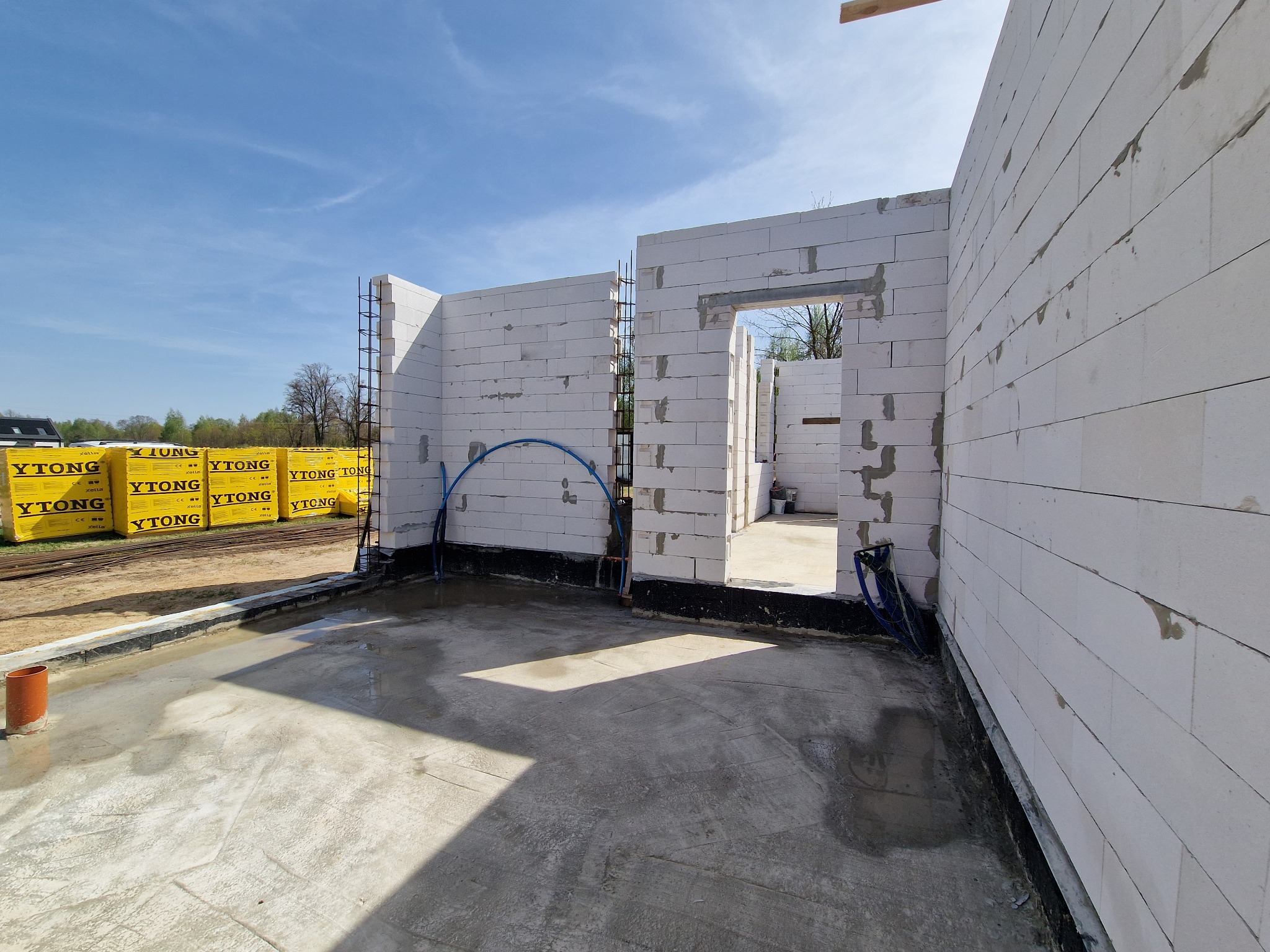 Wnętrze budowanego domu z bloczków Ytong, widoczne ściany w trakcie wznoszenia, betonowa posadzka i przygotowane materiały budowlane na zewnątrz.