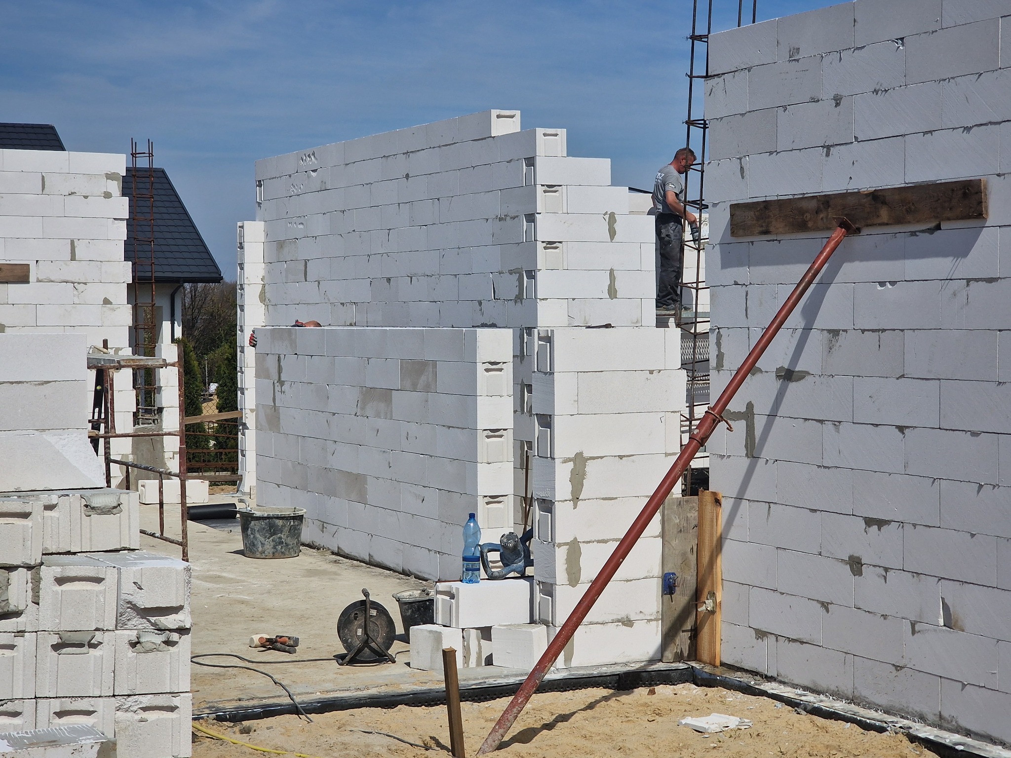 Wznoszenie ścian domu z bloczków Ytong w Łodzi; widoczny murarz na rusztowaniu, podpora, bloczki, wiadro, przedłużacz i butelka wody.