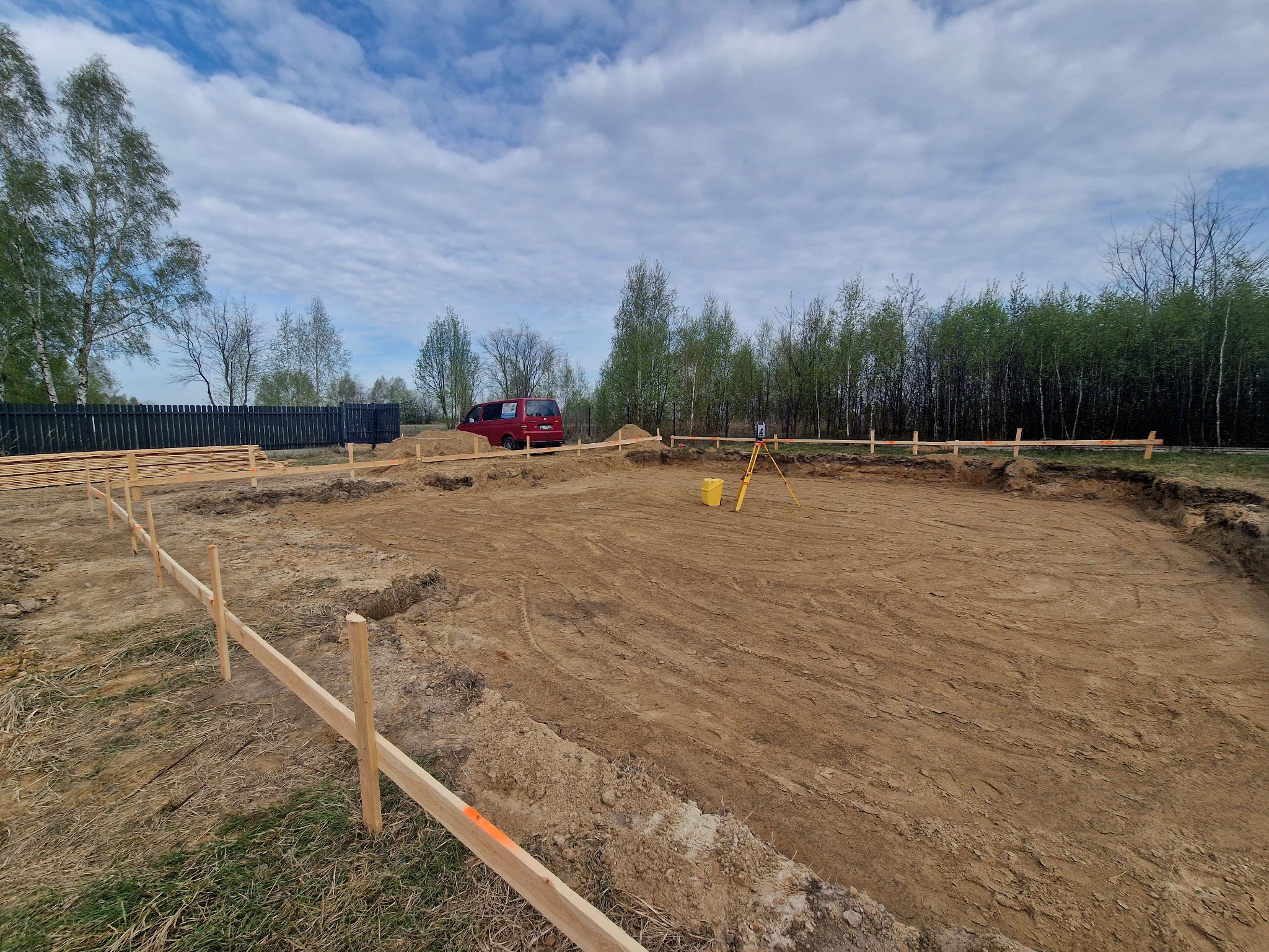 Wytyczony teren pod budowę domu z widocznymi deskami szalunkowymi, niwelatorem na trójnogu i zaparkowanym czerwonym busem w tle.