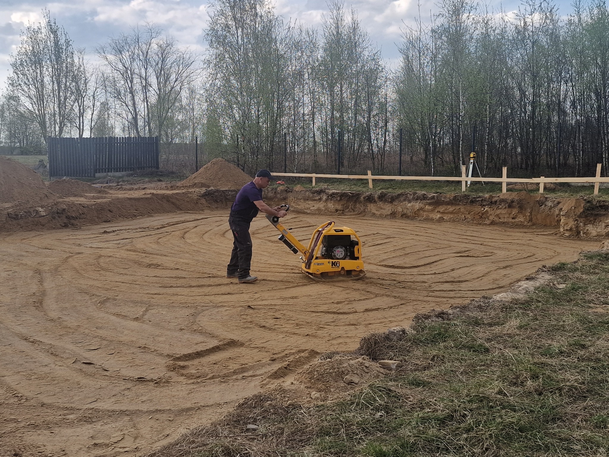 Pracownik zagęszcza piasek zagęszczarką wibracyjną na przygotowanym terenie pod budowę, widoczne ślady po przejazdach maszyny, w tle drzewa i ogrodzenie.