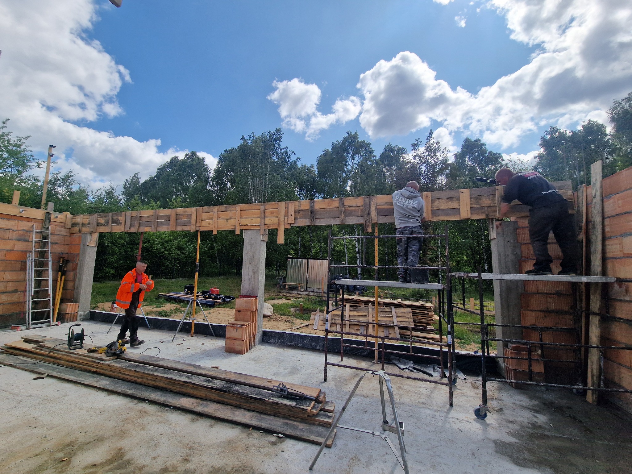 Budowa domu z cegły: murarze montują drewniane szalunki na ścianach, widoczne rusztowania, narzędzia i materiały budowlane na placu budowy w słoneczny dzień.