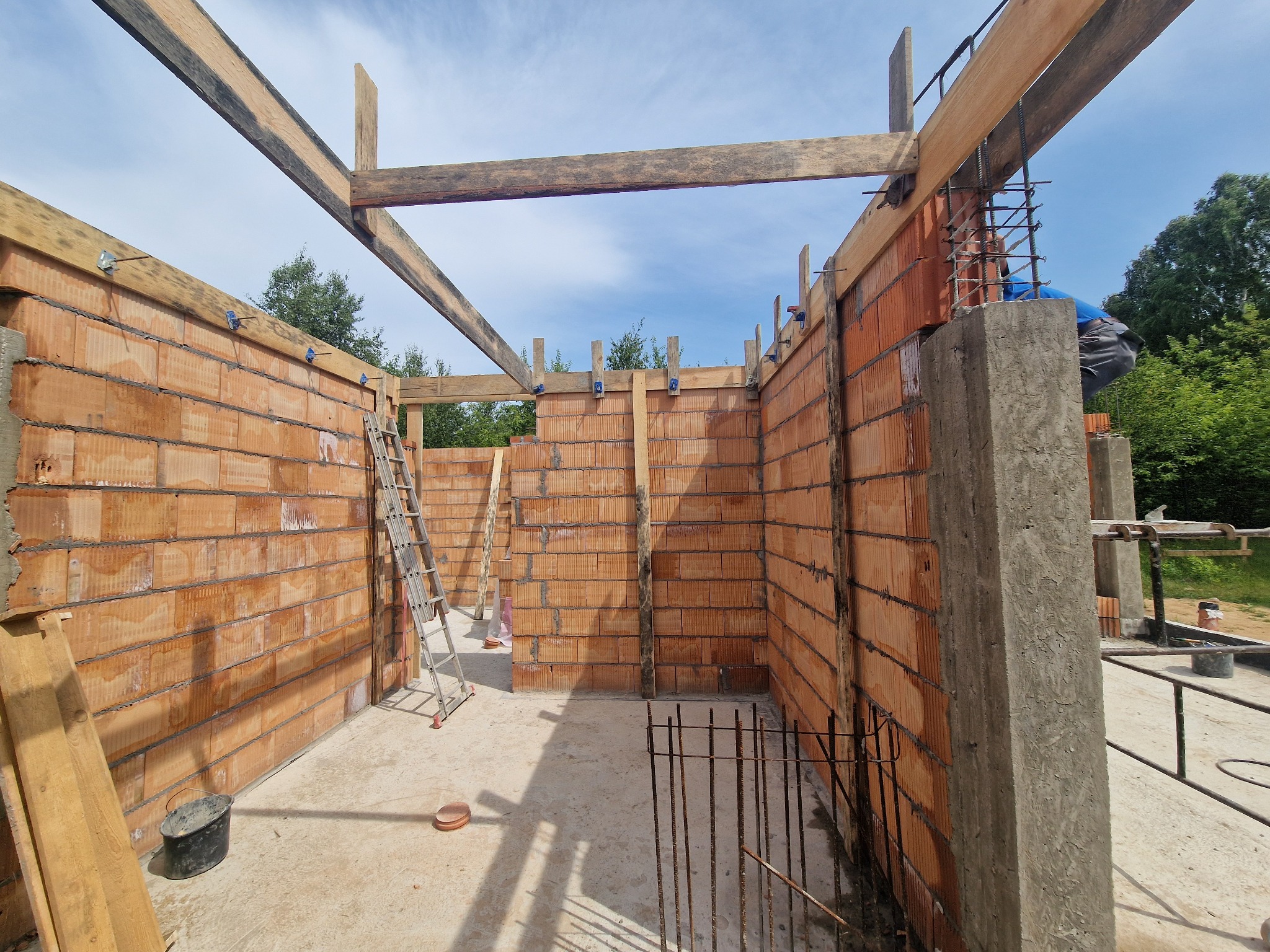 Wznoszenie ścian z pustaków ceramicznych na budowie domu, widoczne zbrojenia i drewniana konstrukcja stropu. Na pierwszym planie betonowa podstawa kolumny.