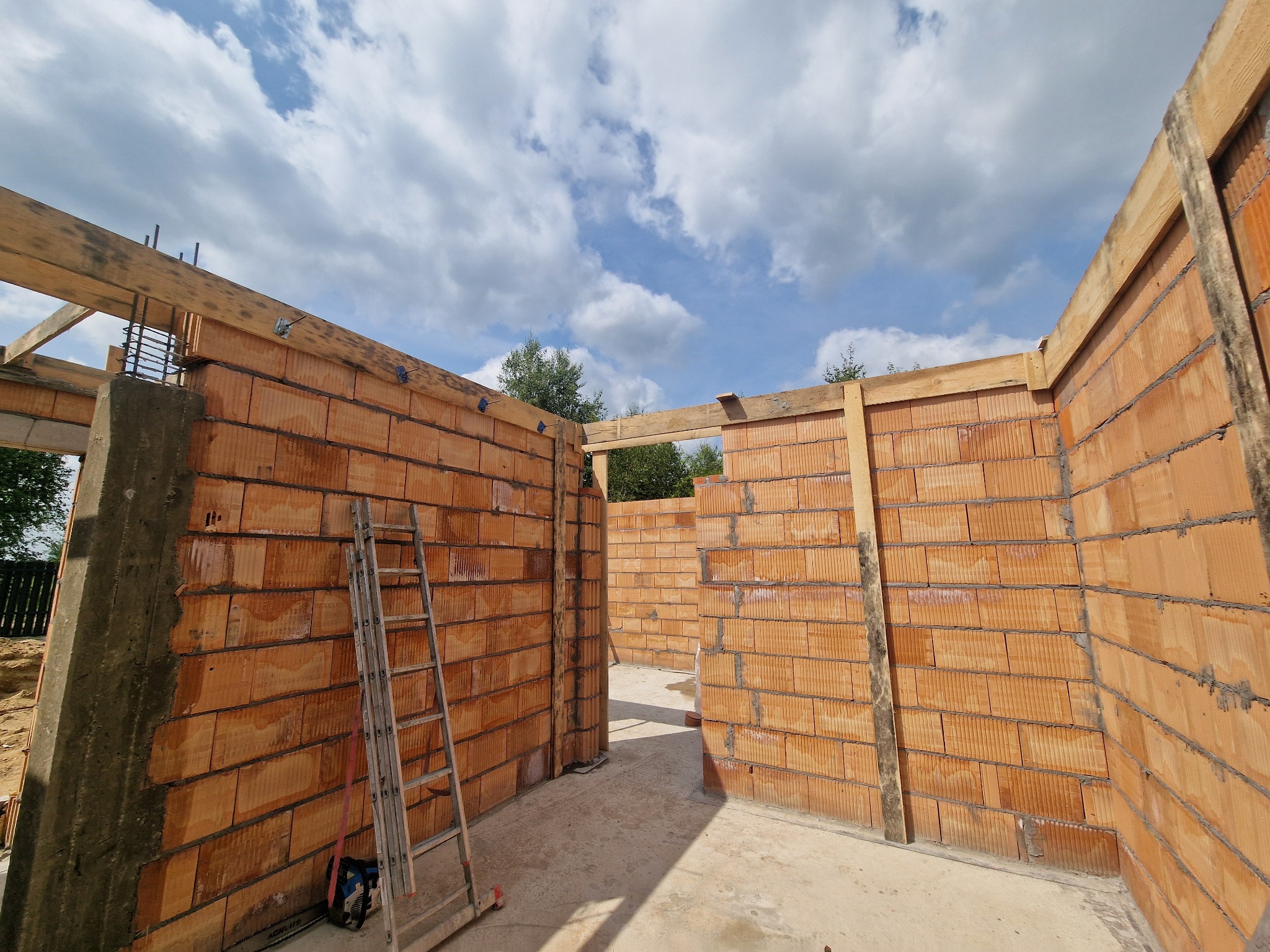 Wnętrze domu w trakcie budowy z cegły ceramicznej, widoczne ściany, drabina oparta o ścianę, betonowy fundament i drewniane elementy konstrukcyjne pod dachem.
