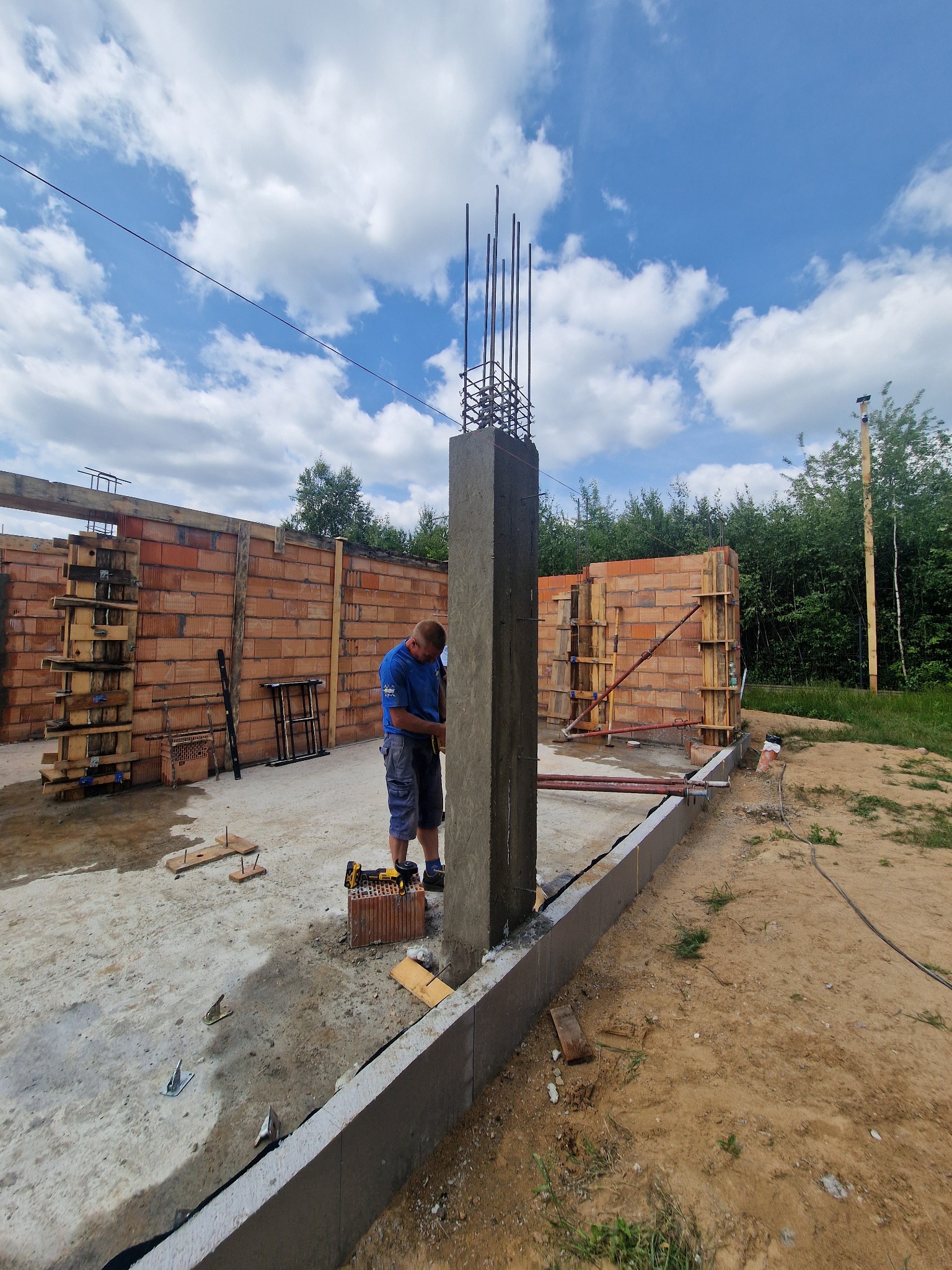 Prace przy wznoszeniu zbrojonego słupa na fundamencie domu z czerwonej cegły. Widoczny pracownik w niebieskiej koszulce i narzędzia.