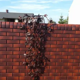 Dekoracyjny, metalowy motyw winogron na ceglanym murze ogrodzeniowym, z widocznym fragmentem budynku w tle.