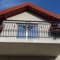 Czarna, metalowa balustrada balkonowa z ozdobnymi elementami, widok z dołu, na tle jasnej elewacji i błękitnego nieba.