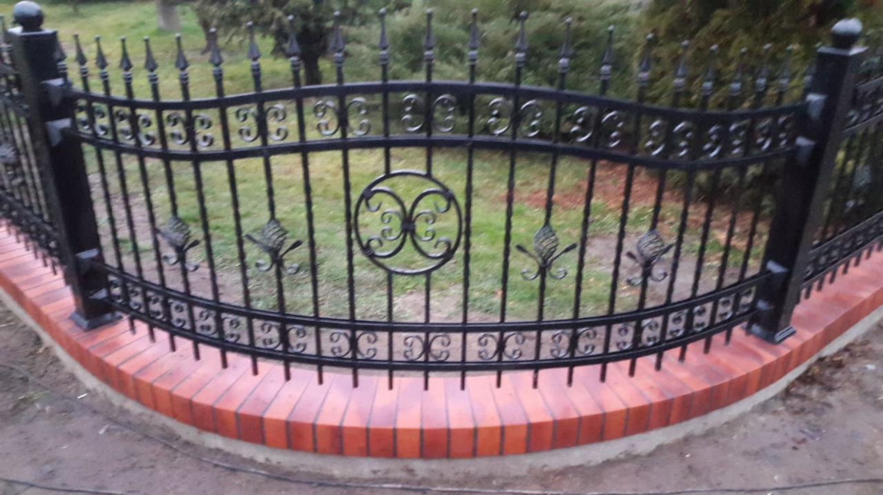 Kute, czarne ogrodzenie z dekoracyjnymi elementami liści i okręgów, osadzone na niskim murku z czerwonej cegły. Widok z bliska, trawnik w tle.