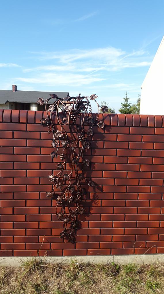 Dekoracyjny, metalowy motyw winogron na ceglanym murze ogrodzeniowym, z widocznym fragmentem budynku w tle.