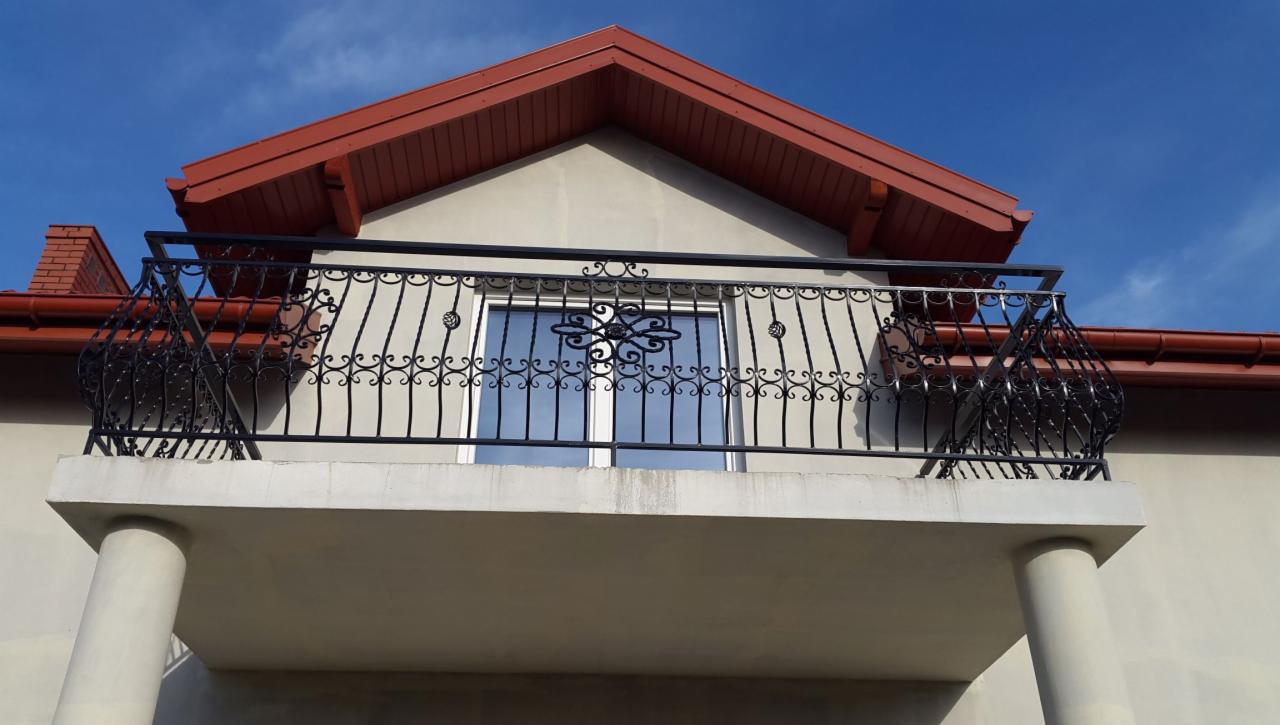 Czarna, metalowa balustrada balkonowa z ozdobnymi elementami, widok z dołu, na tle jasnej elewacji i błękitnego nieba.