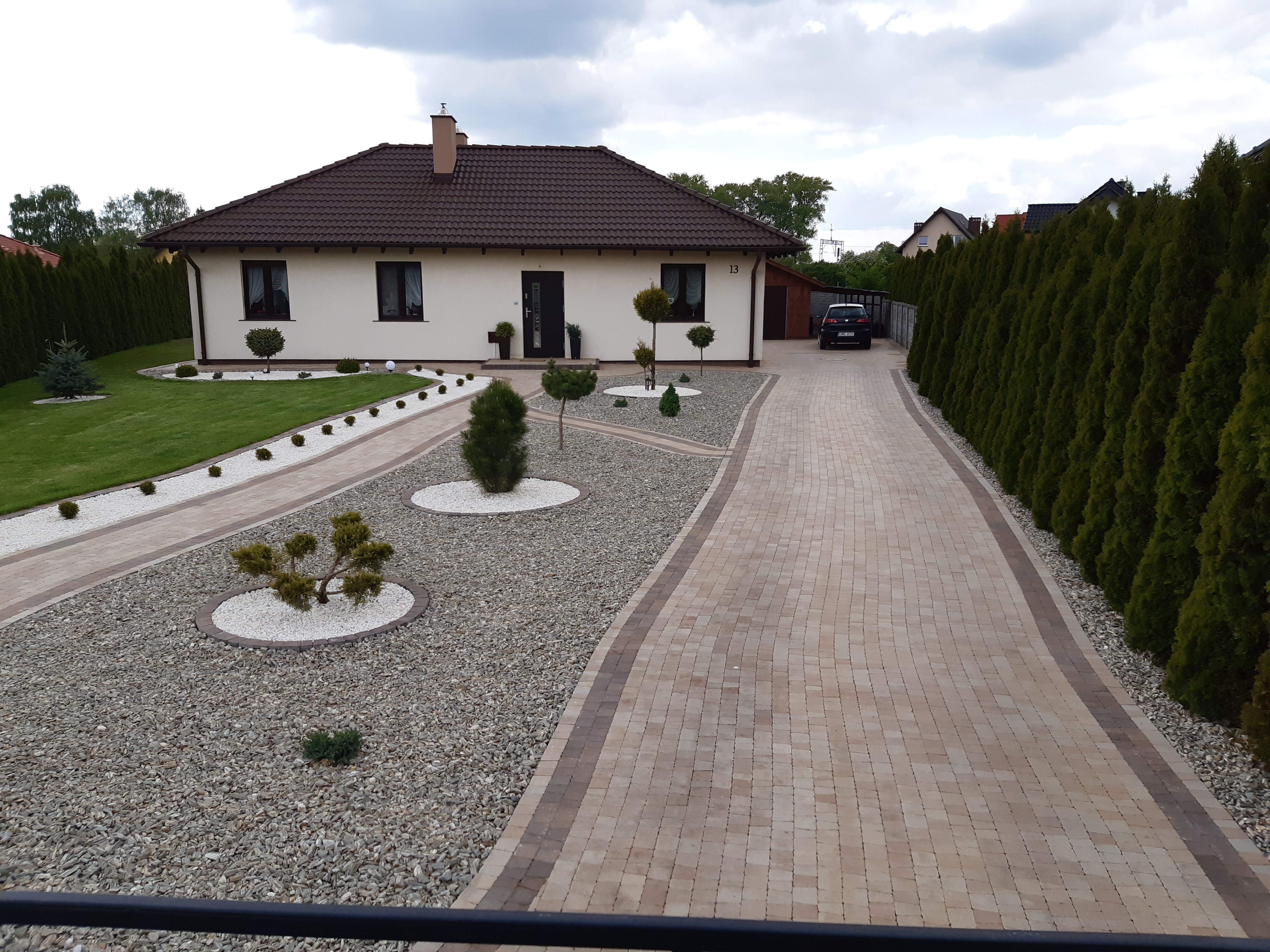 Nowoczesny dom jednorodzinny z jasną elewacją, brązowym dachem i starannie zaprojektowanym ogrodem z ścieżkami z kostki brukowej, ozdobnymi kamieniami i przyciętymi krzewami.