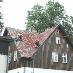 Prace wysokościowe w Bydgoszczy: Dwóch pracowników zabezpieczonych linami na spadzistym, zardzewiałym dachu podczas prac na wysokości. Widoczne okna i fragmenty elewacji budynku.