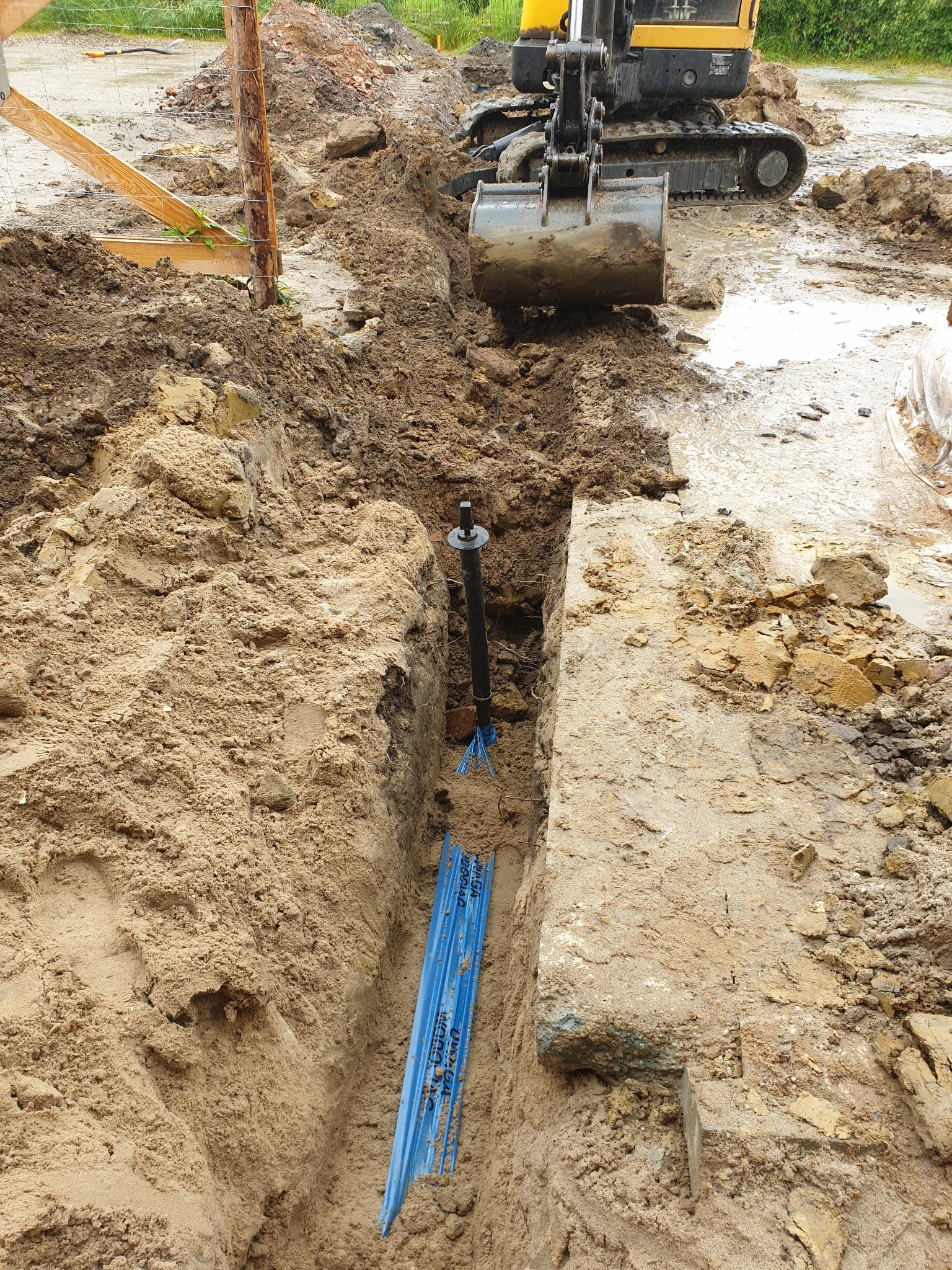 Wykop pod przyłącze wodno-kanalizacyjne z widocznymi niebieskimi rurami, czarną rurą pionową i koparką w tle na grząskim terenie.