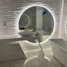 Nowoczesna łazienka z owalnym lustrem podświetlanym LED, biała umywalka nablatowa, bateria w kolorze złota, biała wanna wolnostojąca i dekoracyjne panele ścienne 3D z subtelnym oświetleniem.