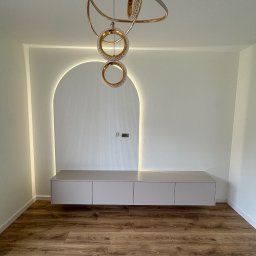 Mk meble na wymiar - Nowoczesna szafka RTV w salonie z podświetlaną wnęką i designerską lampą. Minimalistyczny design, drewniana podłoga, jasne ściany.