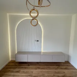 Nowoczesna szafka RTV w salonie z podświetlaną wnęką i designerską lampą. Minimalistyczny design, drewniana podłoga, jasne ściany.