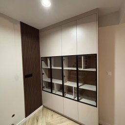 Mk meble na wymiar - Jasna szafa wnękowa z przeszklonymi witrynami i czarnymi ramami, wkomponowana w nowoczesne wnętrze z drewnianą ścianą i parkietem w jodełkę.