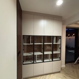 Mk meble na wymiar - Jasna szafa z przeszklonymi drzwiami i czarnymi ramami, wkomponowana we wnętrze z drewnianą ścianą i parkietem w jodełkę. Nowoczesny design, minimalistyczny styl.