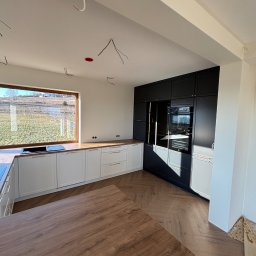 Mk meble na wymiar - Nowoczesna kuchnia z białymi szafkami dolnymi i czarnymi górnymi, drewniany blat i podłoga w jodełkę. Widoczne niedokończone instalacje elektryczne.