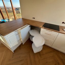 Mk meble na wymiar - Biała kuchnia z drewnianym blatem i wysuwaną półką narożną, widok z góry na parkiet w jodełkę. Duże okno z widokiem na krajobraz.