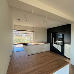 Mk meble na wymiar - Nowoczesna kuchnia z białymi szafkami dolnymi i czarnymi górnymi, drewniany blat, duże okno z widokiem na pole. Wnętrze w trakcie wykończenia, widoczne kable.