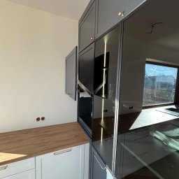 Mk meble na wymiar - Szara zabudowa kuchenna z drewnianym blatem i szklanymi frontami szafek, widoczne otwory na kable w ścianie, złote uchwyty, nowoczesny design.