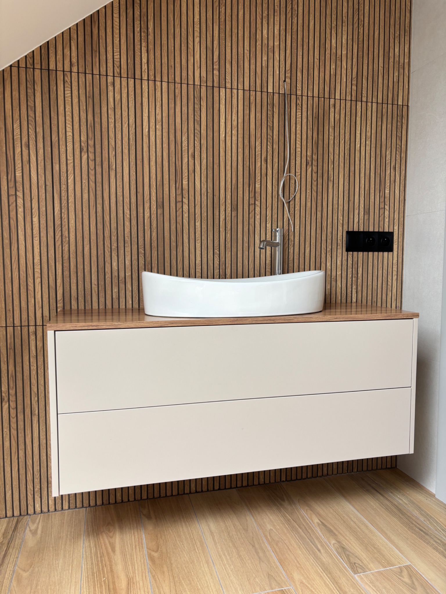 Nowoczesna szafka łazienkowa z umywalką nablatową na tle drewnianej ściany z lameli. Minimalistyczny design, beżowe fronty, drewniany blat.