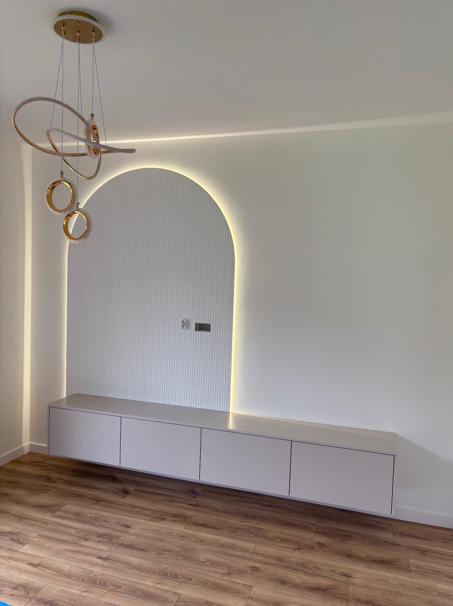 Biała szafka RTV podwieszana na tle ściany z dekoracyjnym, podświetlanym łukiem. Nad nią designerska lampa wisząca. Całość w nowoczesnym stylu.