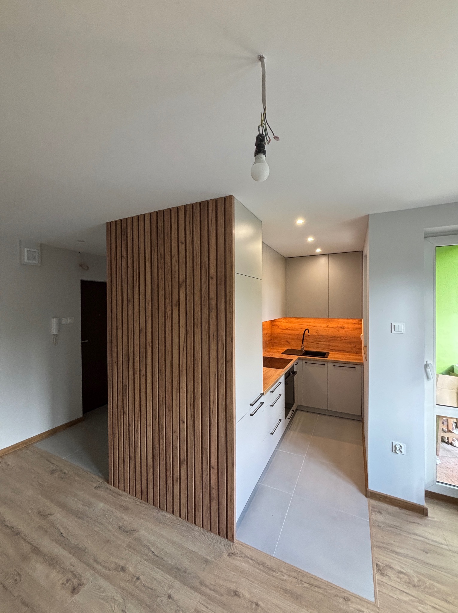 Nowoczesna kuchnia z drewnianym panelem oddzielającym przestrzeń, jasne szafki, drewniany blat i czarna armatura. Widok na drzwi wejściowe i okno.