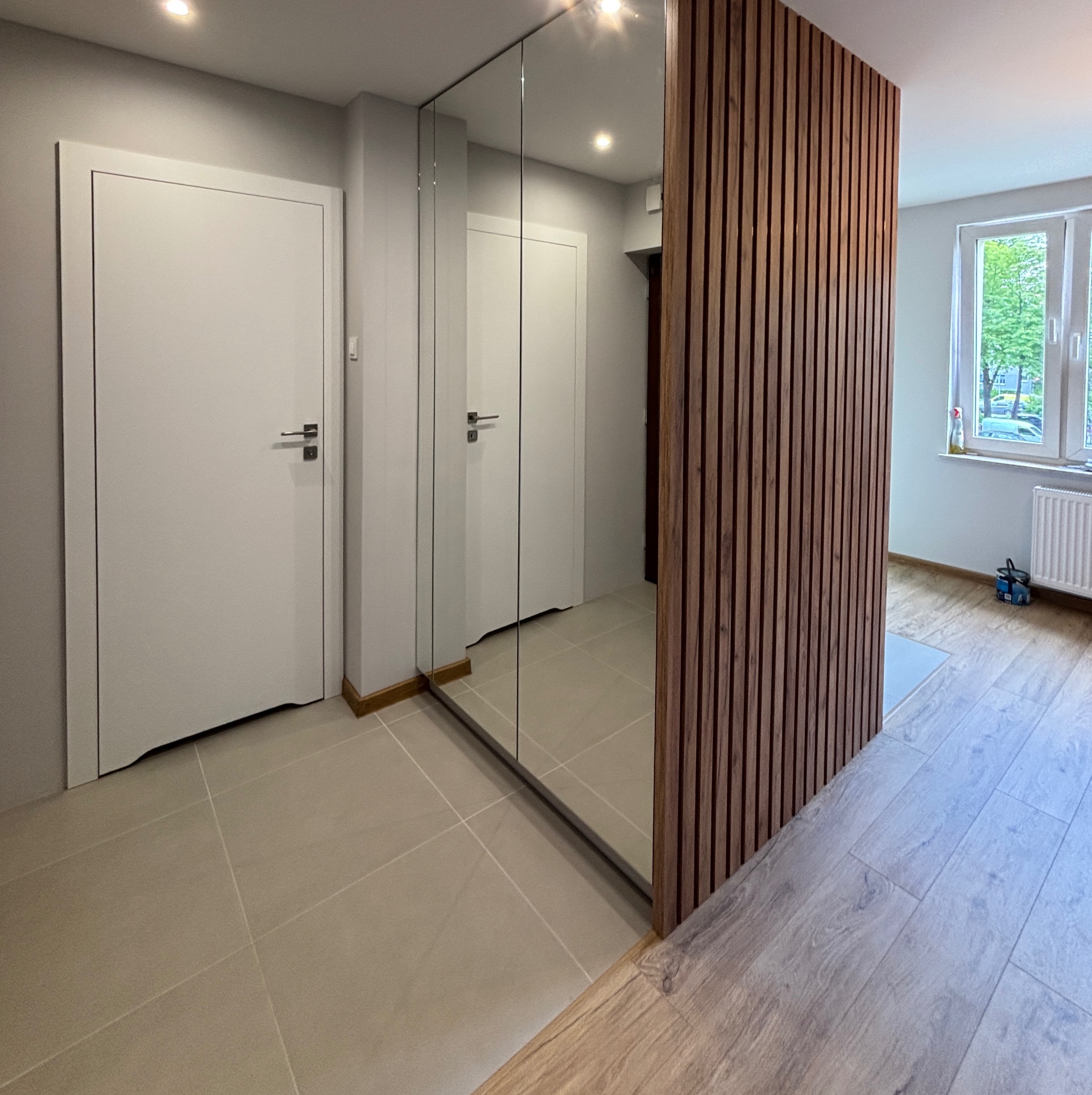 Nowoczesny przedpokój z lustrzaną szafą wnękową i dekoracyjną ścianką z drewnianych listew. Widoczne drzwi i podłoga w odcieniach beżu i szarości.