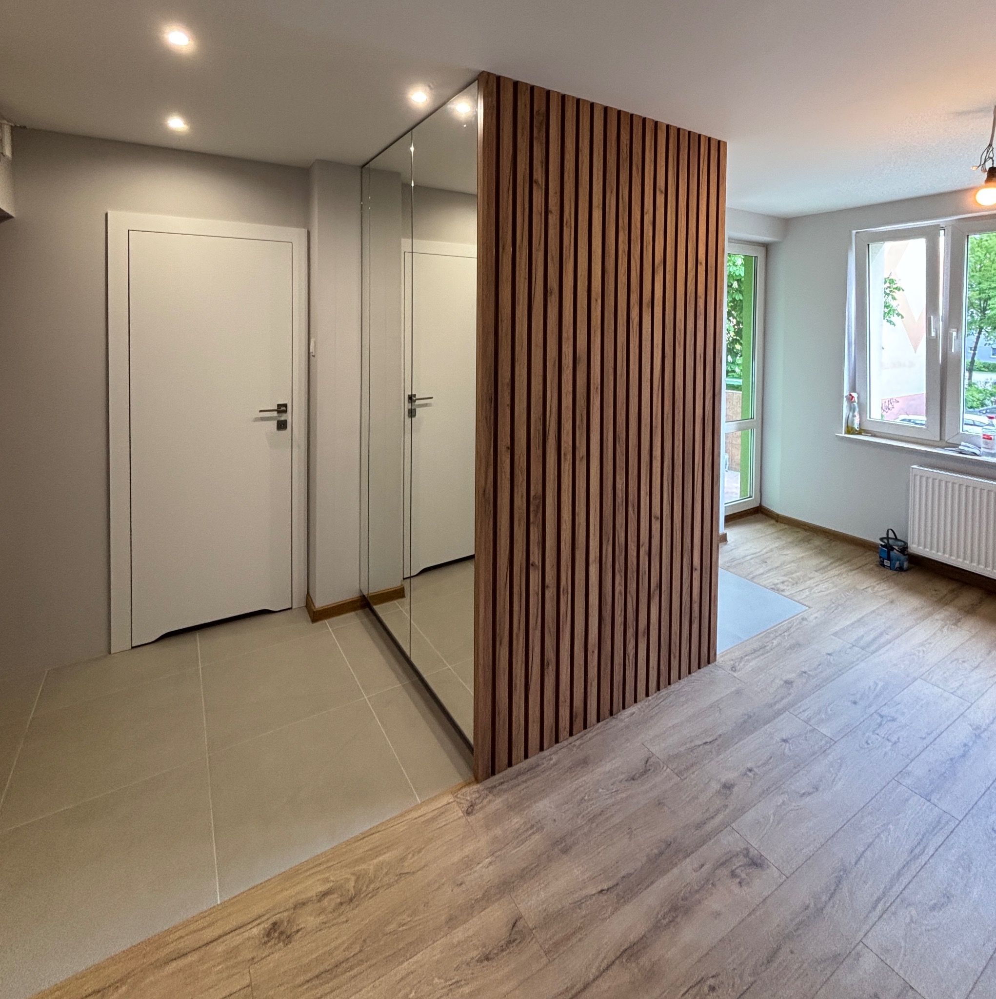 Nowoczesna zabudowa przedpokoju z lustrem i drewnianymi lamelami. Jasne płytki podłogowe i drewnopodobne panele. Minimalistyczny design, oświetlenie punktowe.