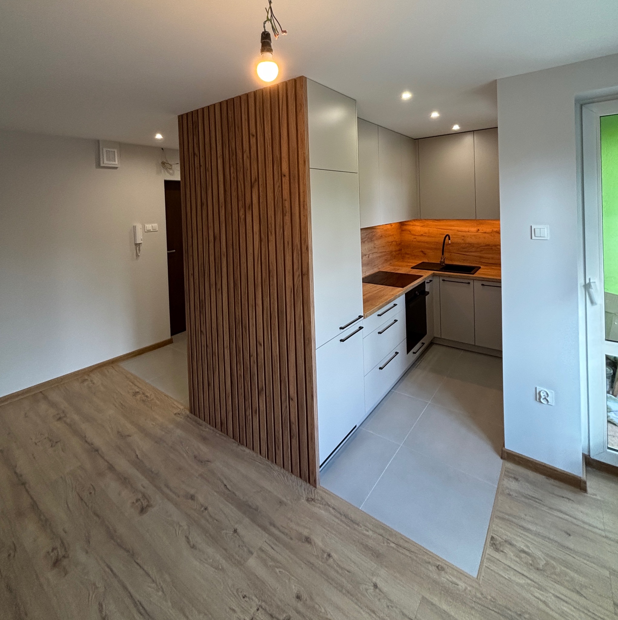 Nowoczesna kuchnia z szarymi szafkami i drewnianym blatem, oddzielona od salonu drewnianą ścianką z lameli. Widoczny fragment podłogi i wisząca żarówka.