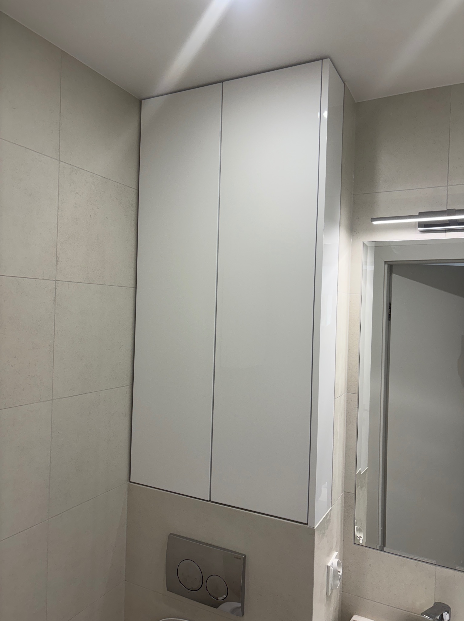 Biała szafka łazienkowa na wymiar z połyskiem, zamontowana nad toaletą, z widocznym przyciskiem spłukiwania i fragmentem lustra. Minimalistyczny design.