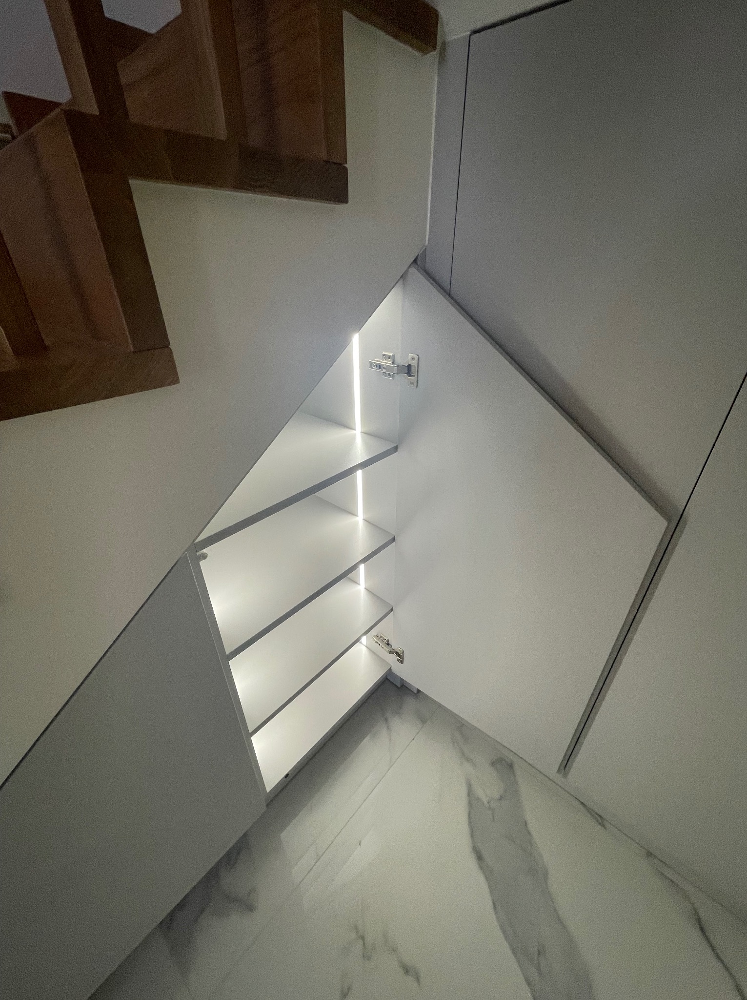 Szafa wnękowa pod schodami z oświetleniem LED, białe fronty, otwarte półki, marmurowa podłoga, widok z dołu, nowoczesny design.