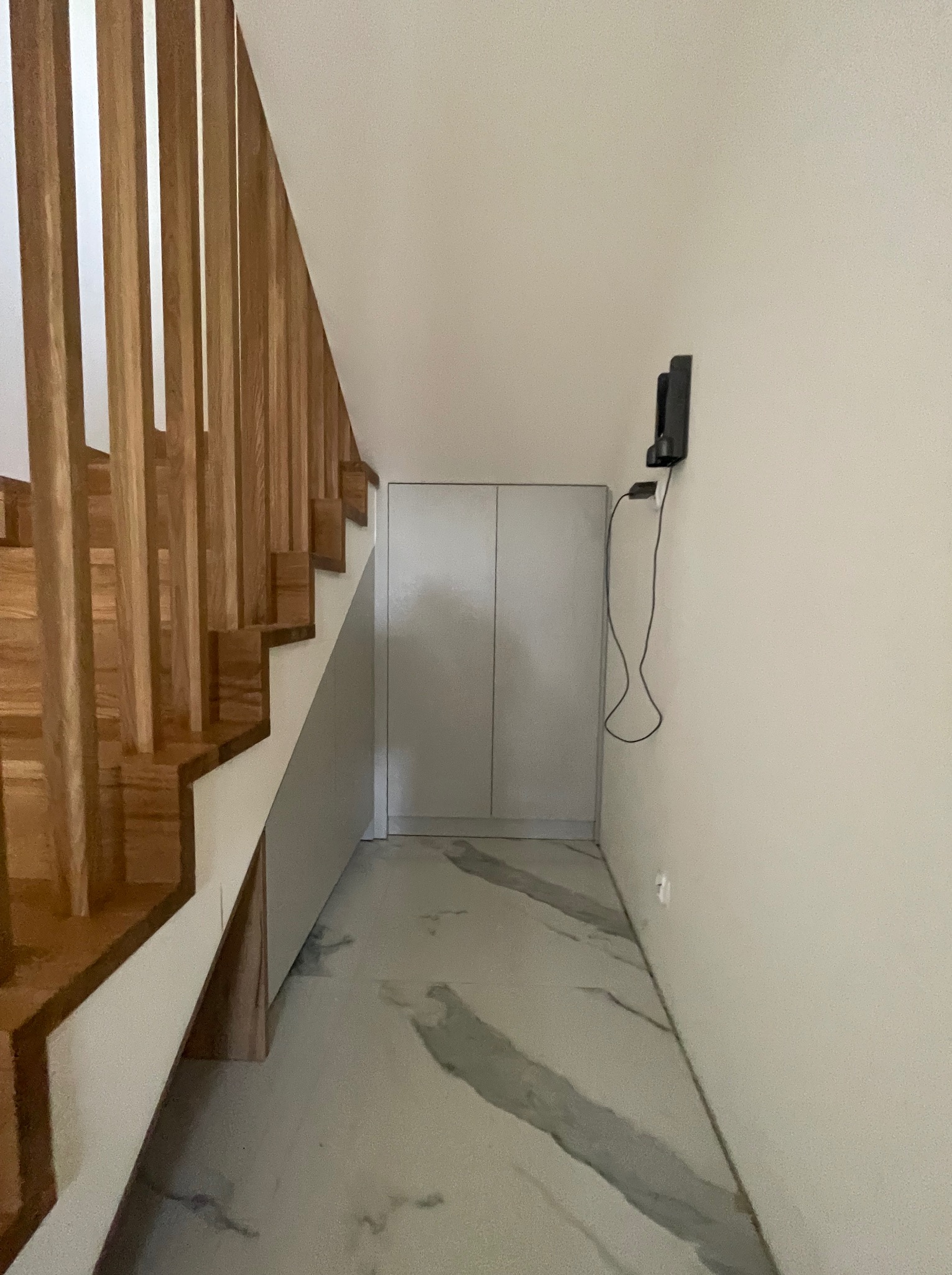 Biała szafa wnękowa pod schodami z drewnianą balustradą, marmurowa podłoga, minimalistyczny design, oświetlenie punktowe na ścianie.