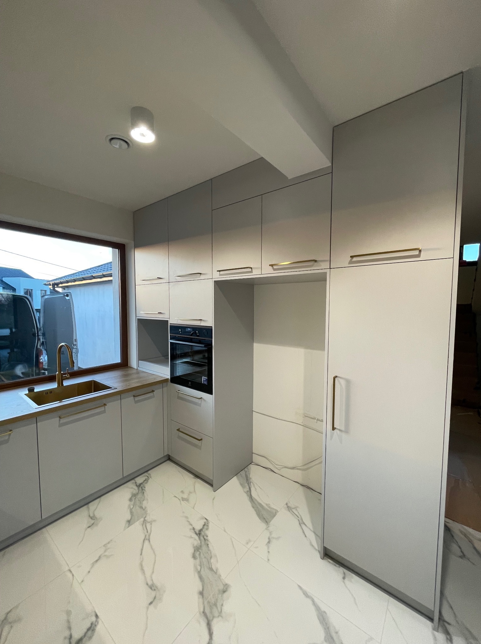 Nowoczesna kuchnia w odcieniach szarości z złotymi akcentami: bateria, uchwyty. Marmurowe płytki podłogowe. Widok na zewnątrz przez okno.