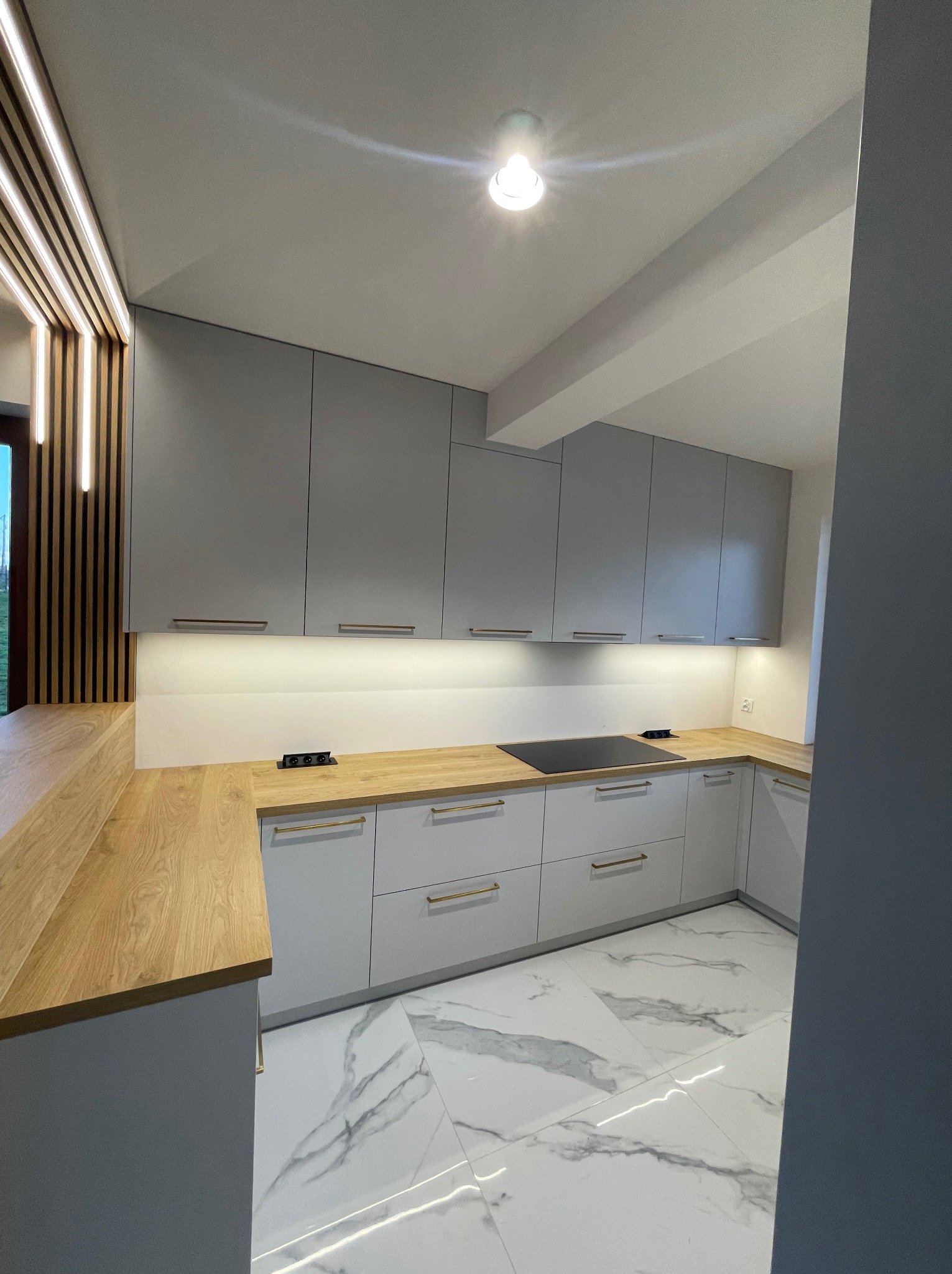 Nowoczesna kuchnia na wymiar: szare szafki z drewnianym blatem i marmurową podłogą. Oświetlenie LED podkreśla minimalistyczny design.