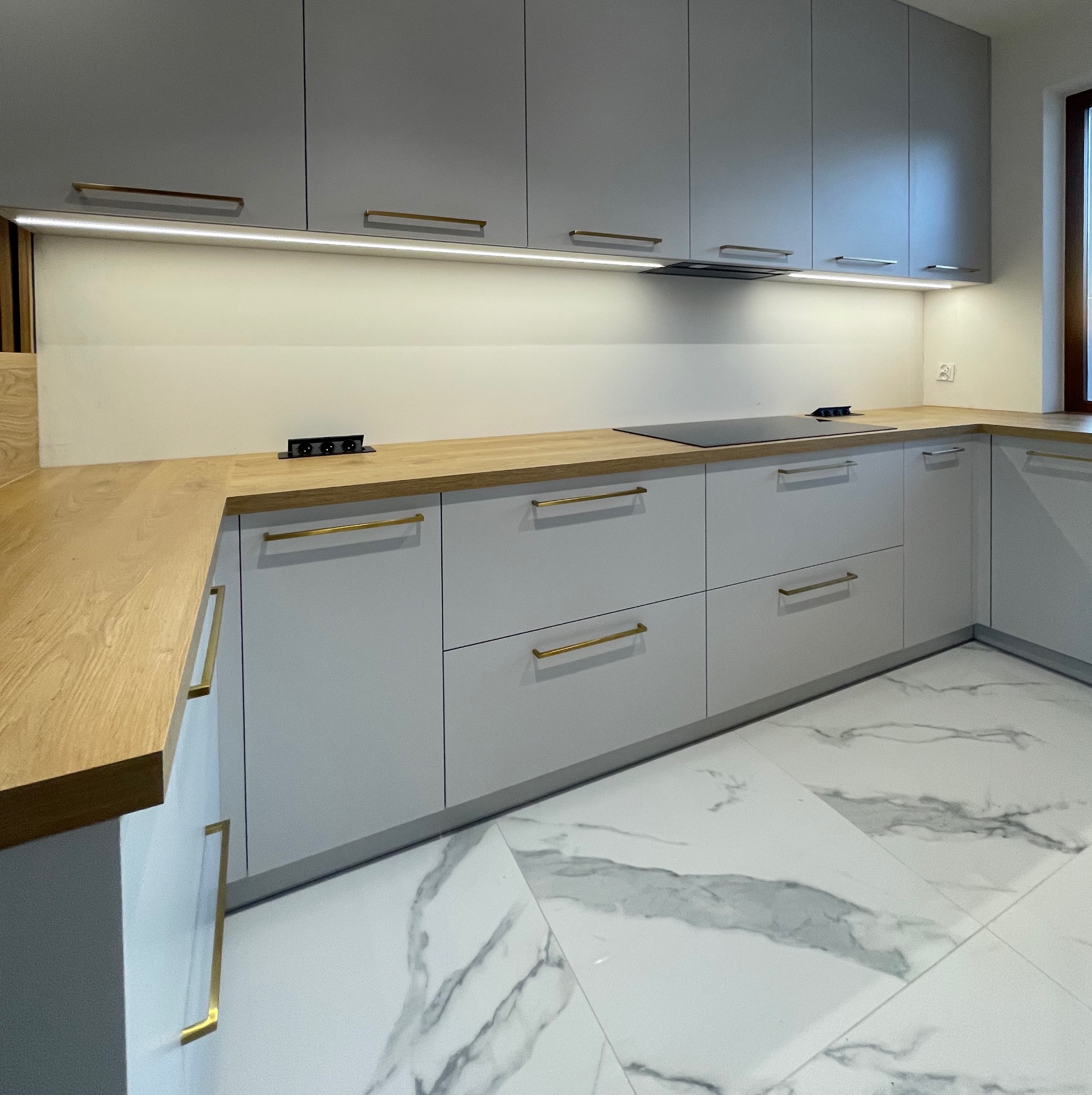 Szara kuchnia z drewnianym blatem i złotymi uchwytami, oświetlenie LED pod szafkami, marmurowe płytki podłogowe. Nowoczesny design, widok ukośny.