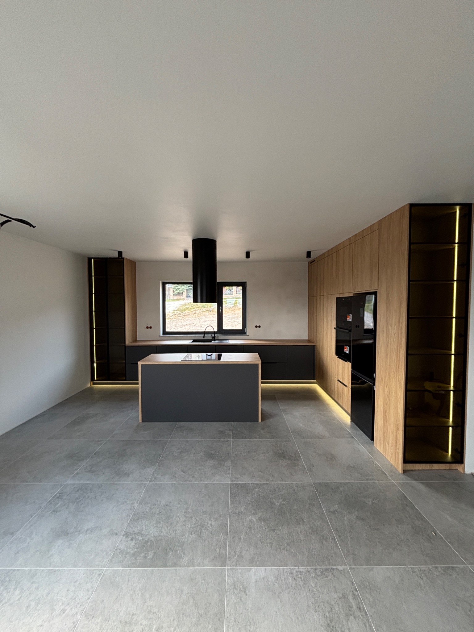 Nowoczesna kuchnia z wyspą, fronty w odcieniach drewna i szarości, oświetlenie LED podkreślające detale. Minimalistyczny design, czarny okap cylindryczny.