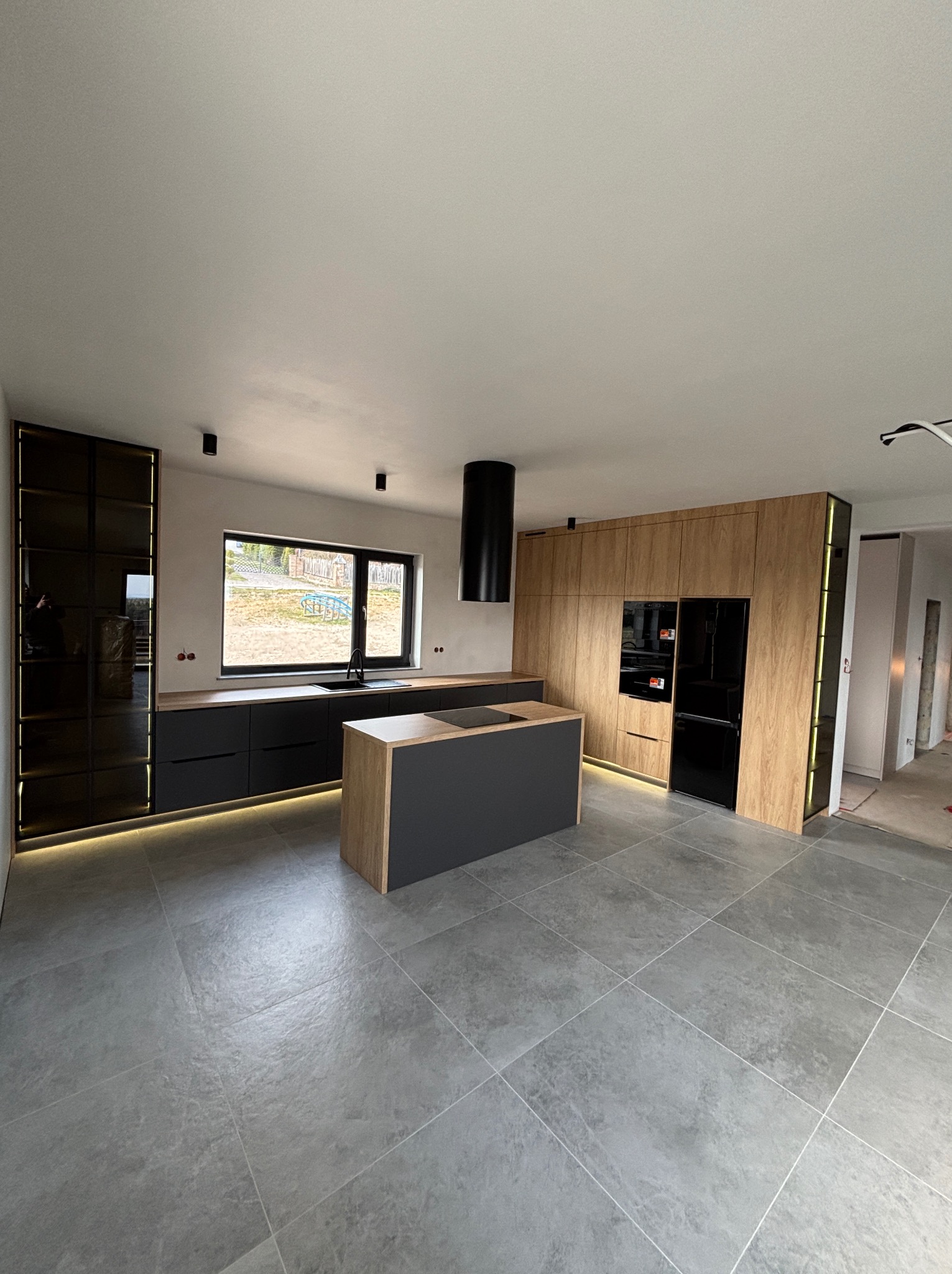 Nowoczesna kuchnia z wyspą: szare fronty, drewniane akcenty, podświetlenie LED. Czarny okap cylindryczny i sprzęt AGD. Duże płytki podłogowe. Minimalistyczny design.