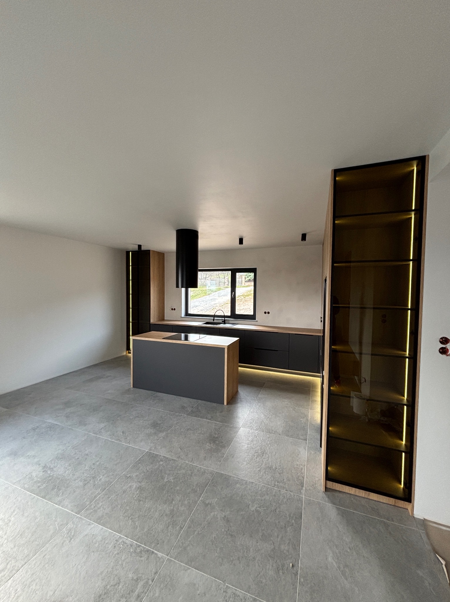 Nowoczesna kuchnia z wyspą i podświetlanymi szafkami. Minimalistyczny design, szare płytki podłogowe, czarne akcenty i drewniane wykończenia. Duże okno z widokiem na zewnątrz.