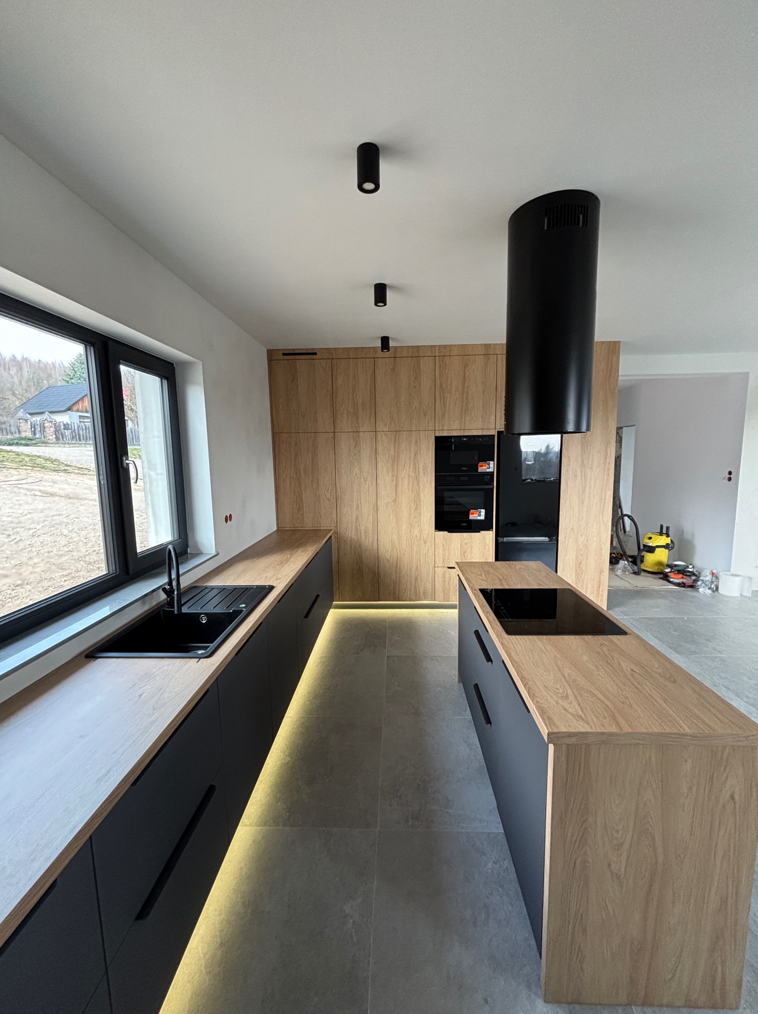 Nowoczesna kuchnia z wyspą: drewniane fronty szafek, czarne blaty i oświetlenie LED pod szafkami. Minimalistyczny design, okno z widokiem na zewnątrz.