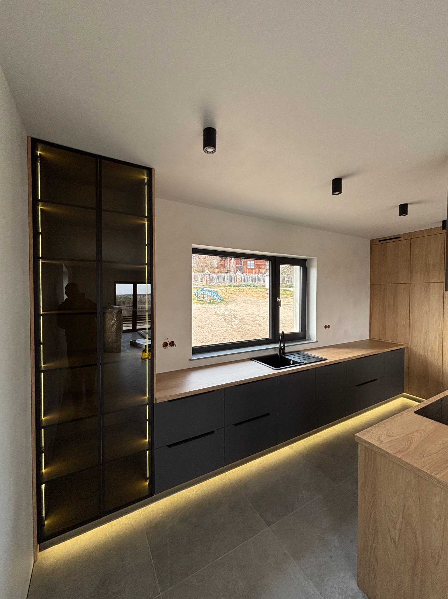 Nowoczesna kuchnia z czarnymi szafkami, drewnianym blatem i podświetleniem LED. Wysoka szafa z przydymionego szkła z oświetleniem wewnętrznym. Minimalistyczny design.