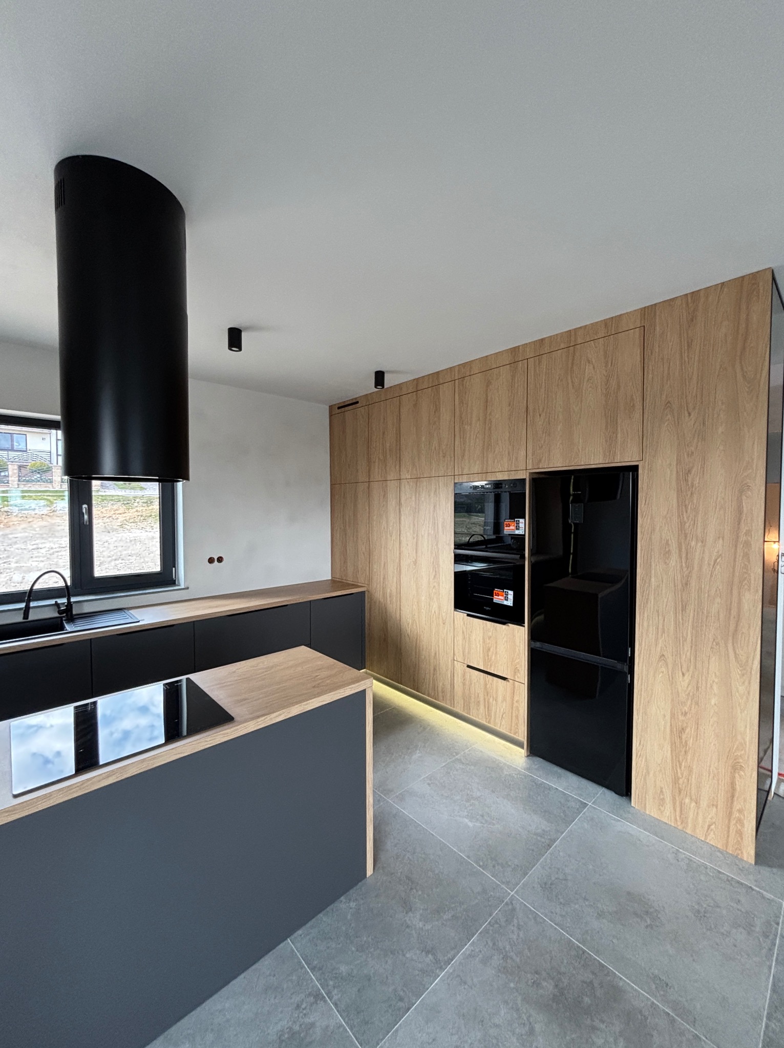 Nowoczesna kuchnia z drewnianymi frontami, czarnymi elementami i wyspą. Minimalistyczny design, oświetlenie LED pod szafkami. Widok na okno i otoczenie.