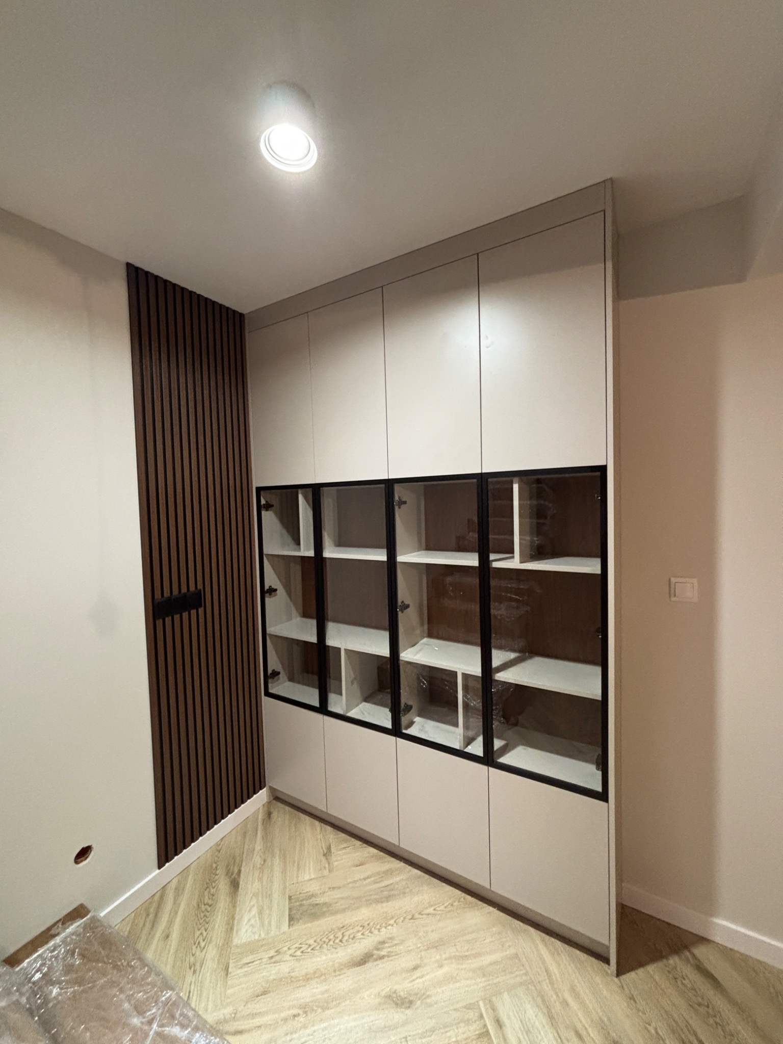 Jasna szafa wnękowa z przeszklonymi witrynami i czarnymi ramami, wkomponowana w nowoczesne wnętrze z drewnianą ścianą i parkietem w jodełkę.